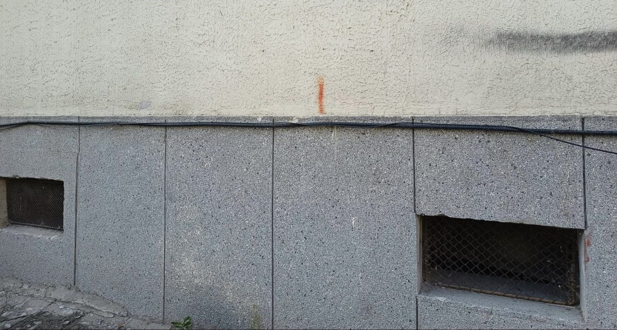Image shows a blank wall. Two grates and grey concrete make up the bottom half of the wall and the top half of the wall is off-white colour. There is one distinctive small vertical red line of paint on the wall where the off-white half meets the grey half.